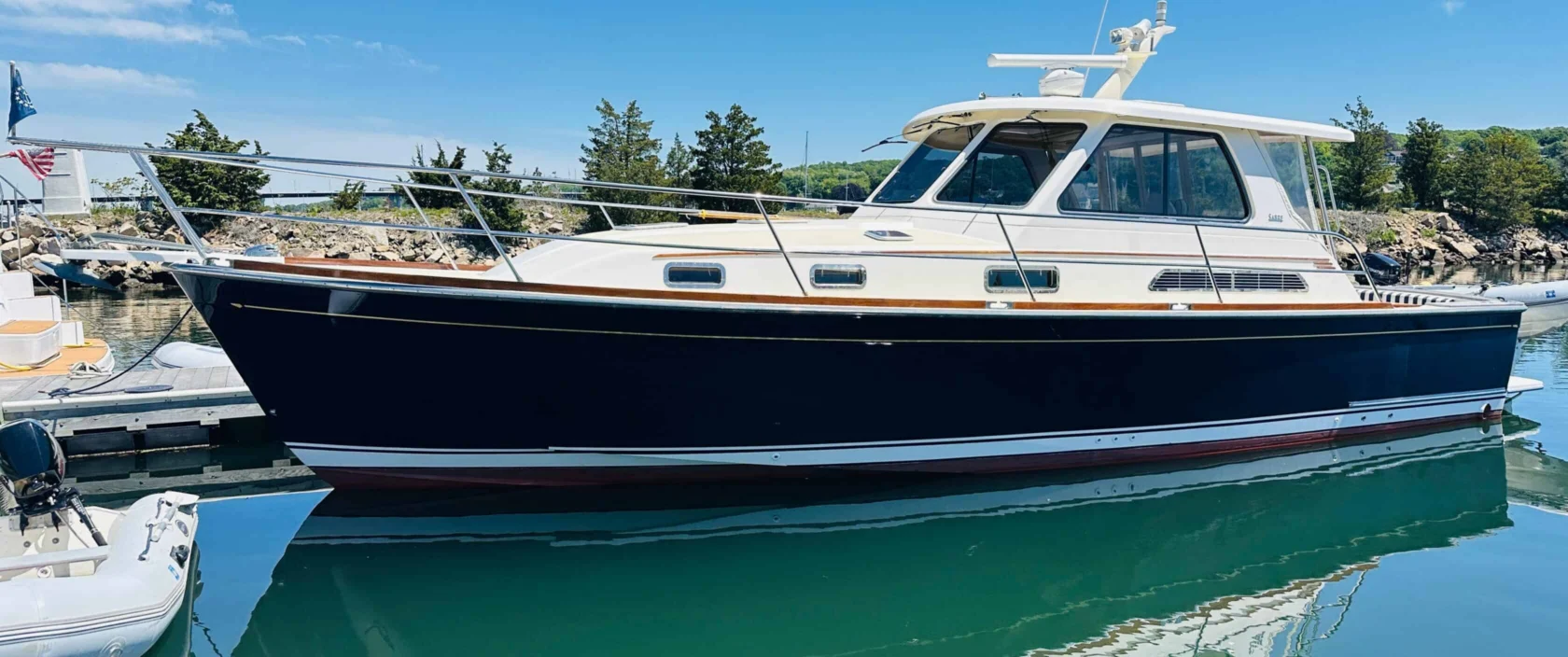 2006 Sabre 38 Hard Top Express yacht Piacere with an Awlgrip flag blue hull docked at a marina, reflecting in calm green water under a blue sky.